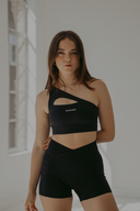 Woman in black one-shoulder sports bra and high-waist shorts posing in a bright studio, minimalist activewear look.