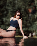 Woman in a black swimsuit and sunglasses sitting by a pool with trees in the background